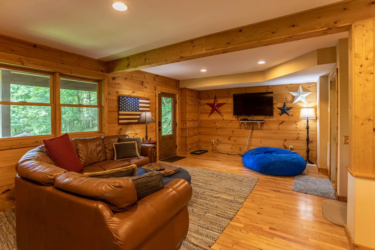A cozy second living area with a leather sofa, American flag wall art, blue bean bag, wooden walls, and a large smart TV. The arrangement is perfect for relaxing, watching movies, or enjoying game nights with friends and family.