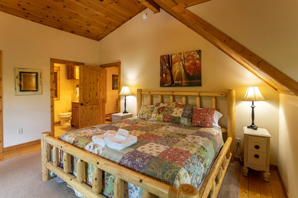 A rustic bedroom with a wooden bed, colorful quilt, framed artwork, and soft bedside lamps. Morning sunlight streams through unseen windows, highlighting the cozy textures and inviting one to relax or read in bed.