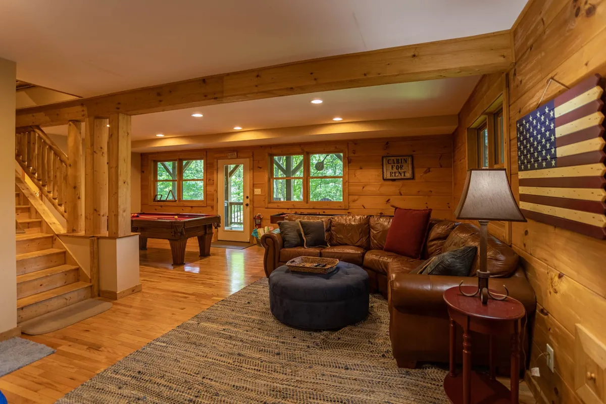 A warm, inviting game room with a pool table, leather sofa, wooden beams, large windows, and American flag décor. The space is designed for fun family nights with views of the outdoors, blending recreation and relaxation after a day of adventures.