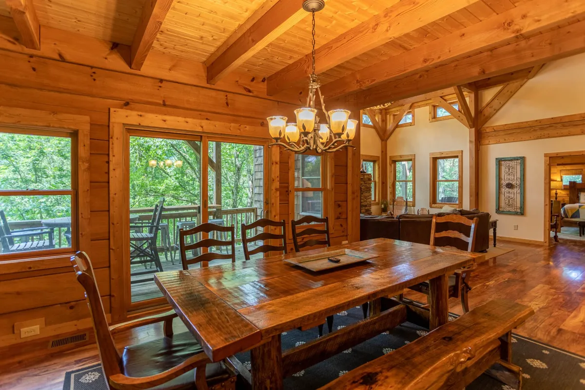 A spacious dining area with a large wooden table, chandelier, and expansive views of the surrounding trees through tall windows. The setup suggests a blend of communal gathering and connection to nature.