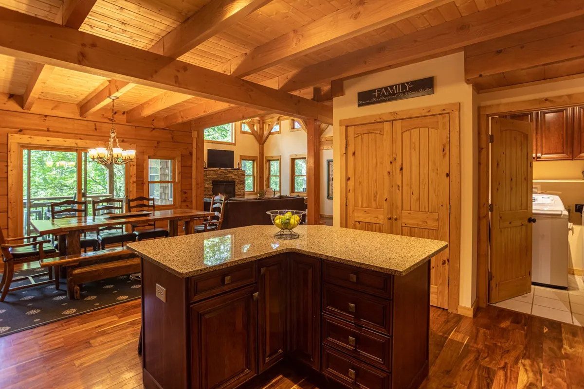 A view connecting the kitchen island, dining table, rustic wooden beams, fireplace, and large windows. The space looks both elegant and comfortable, ideal for hosting family gatherings, cooking, dining, and relaxing by the fire while enjoying the scenery.