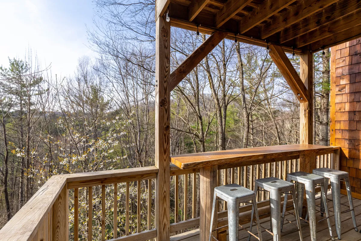 This image captures an inviting outdoor bar counter with metal stools set on a wooden deck, surrounded by lush trees. The scene exudes a relaxed, convivial atmosphere ideal for group gatherings, casual drinks, and enjoying the mountain air.