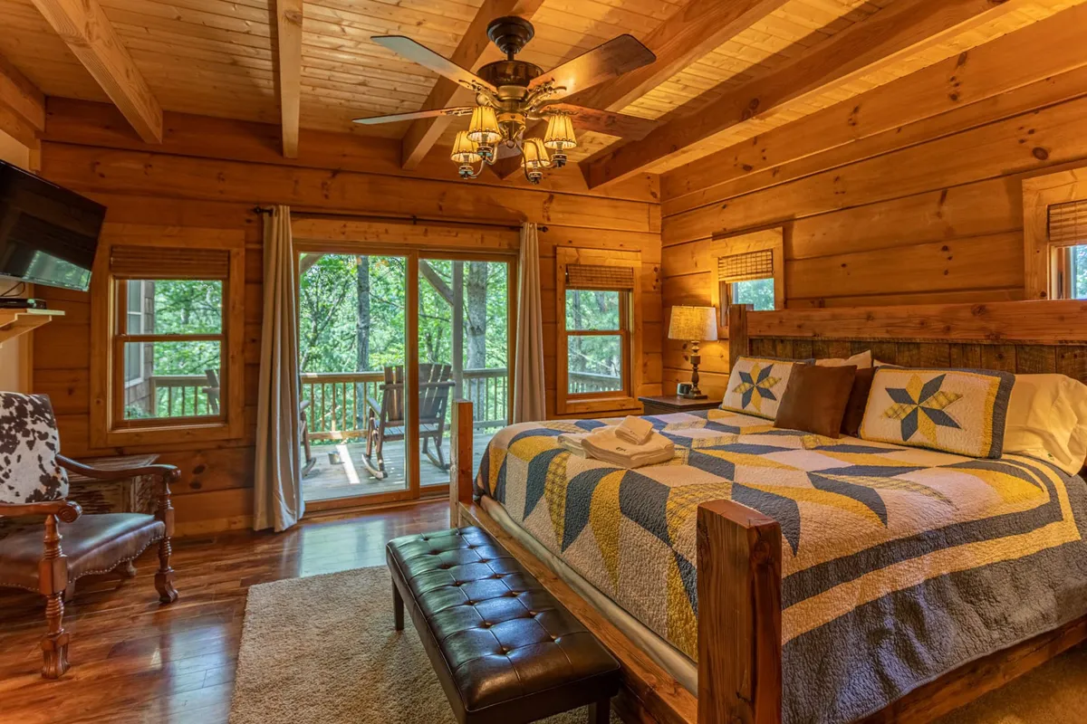 A warm, inviting bedroom with a bed dressed in a colorful quilt, large windows with views of the surrounding nature, a comfortable armchair for reading, wooden ceiling, and a charming light fixture. The space feels serene and ideal for quiet relaxation or enjoying a peaceful morning.