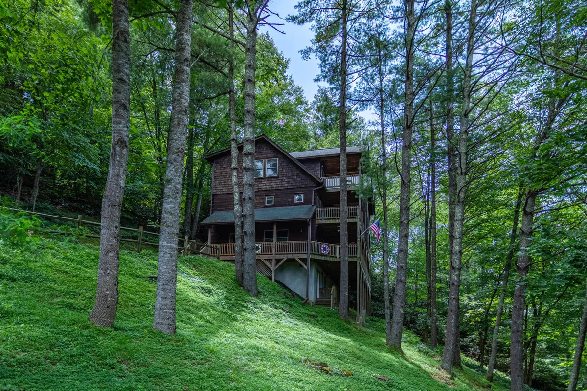 A welcoming view of the wooden house nestled among tall trees, with green grass and an American flag waving near the entrance. The expansive deck suggests space for gatherings and offers a glimpse of the natural surroundings, perfect for family adventures and outdoor activities.