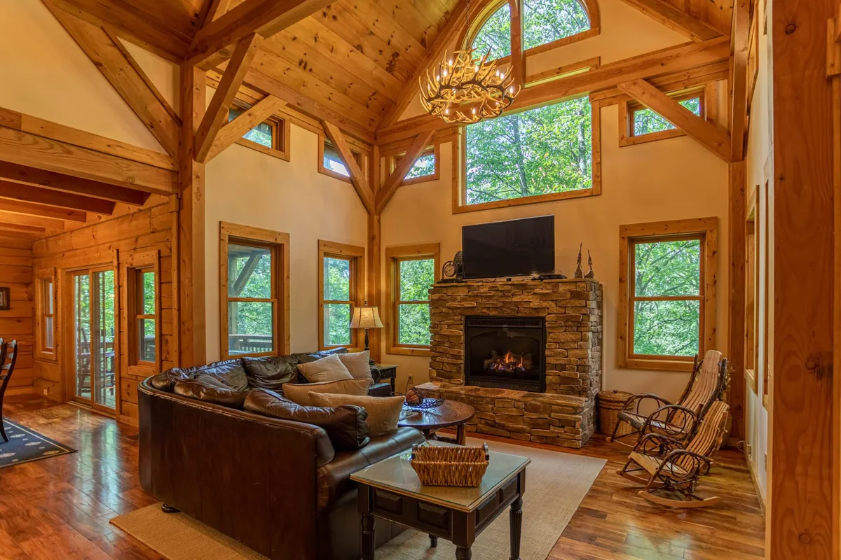 A spacious, inviting living room featuring a majestic stone fireplace as the centerpiece, surrounded by plush seating. Towering wooden beams and expansive windows frame peaceful woodland views, with sunlight filtering through the trees, creating a warm and tranquil ambiance.