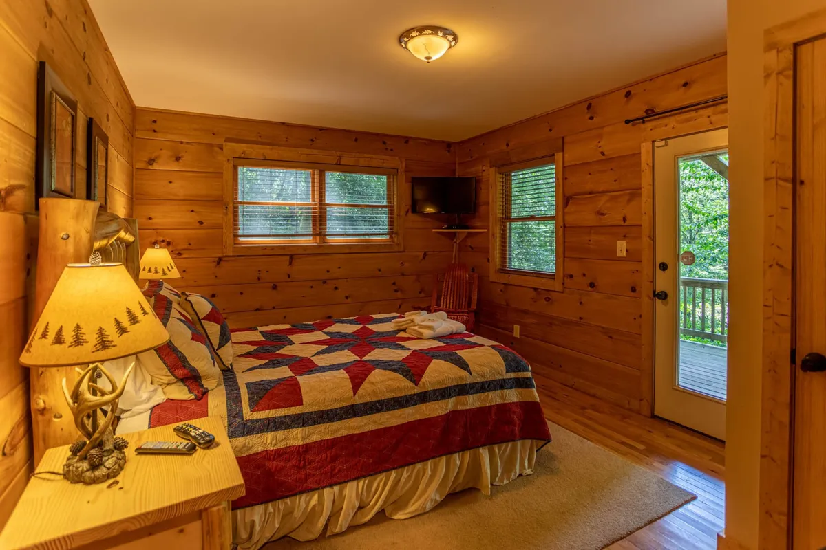 A cozy, rustic bedroom with a quilted bedspread, wooden walls, an armchair, and a door leading outside. Natural light filters in, and the door suggests immediate access to the outdoors, blending interior comfort with the surrounding nature.
