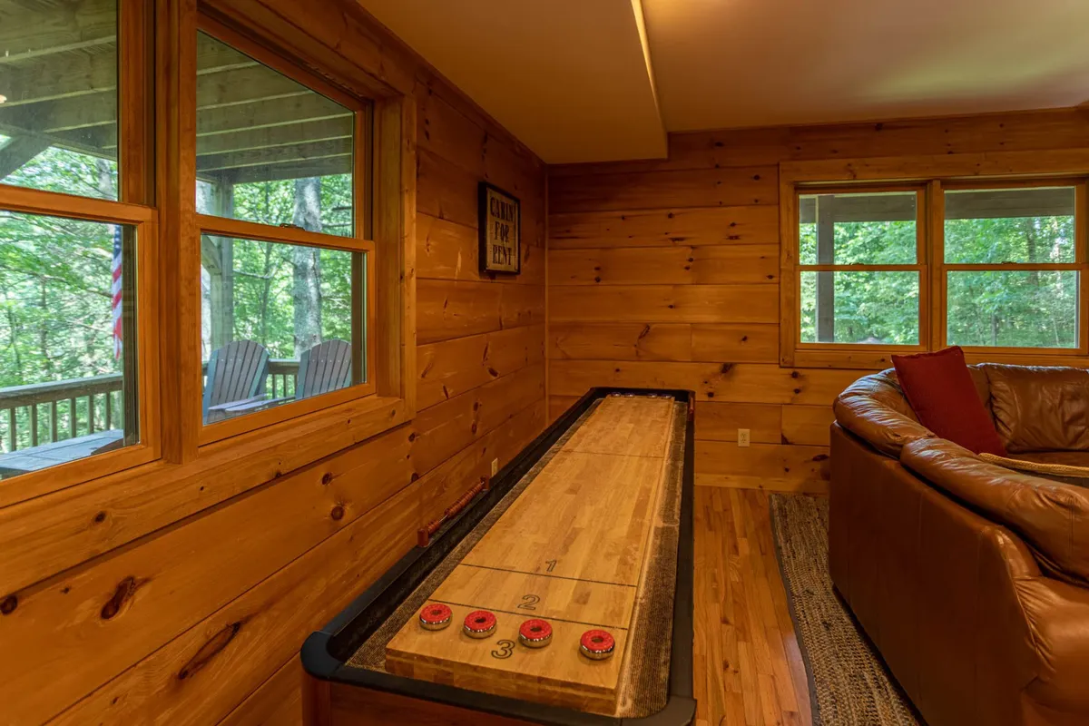 A spacious rec room featuring a shuffleboard table, comfortable seating, and large windows that bring in ample natural light. The wooden walls and overall rustic décor evoke a cozy mountain retreat, while the windows offer glimpses of the outdoor landscape.