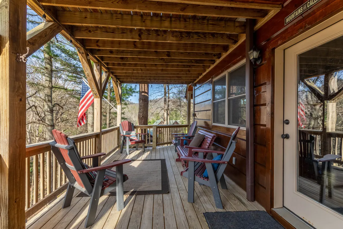 This exterior image showcases a spacious wooden deck outfitted with red chairs and an inviting atmosphere. The deck is fringed by trees, and there's an American flag hanging near the doorway, suggesting a welcoming, homey vibe. The outdoor setting offers a clear connection to nature and plenty of space for relaxing outdoors with pets after a day of adventure.