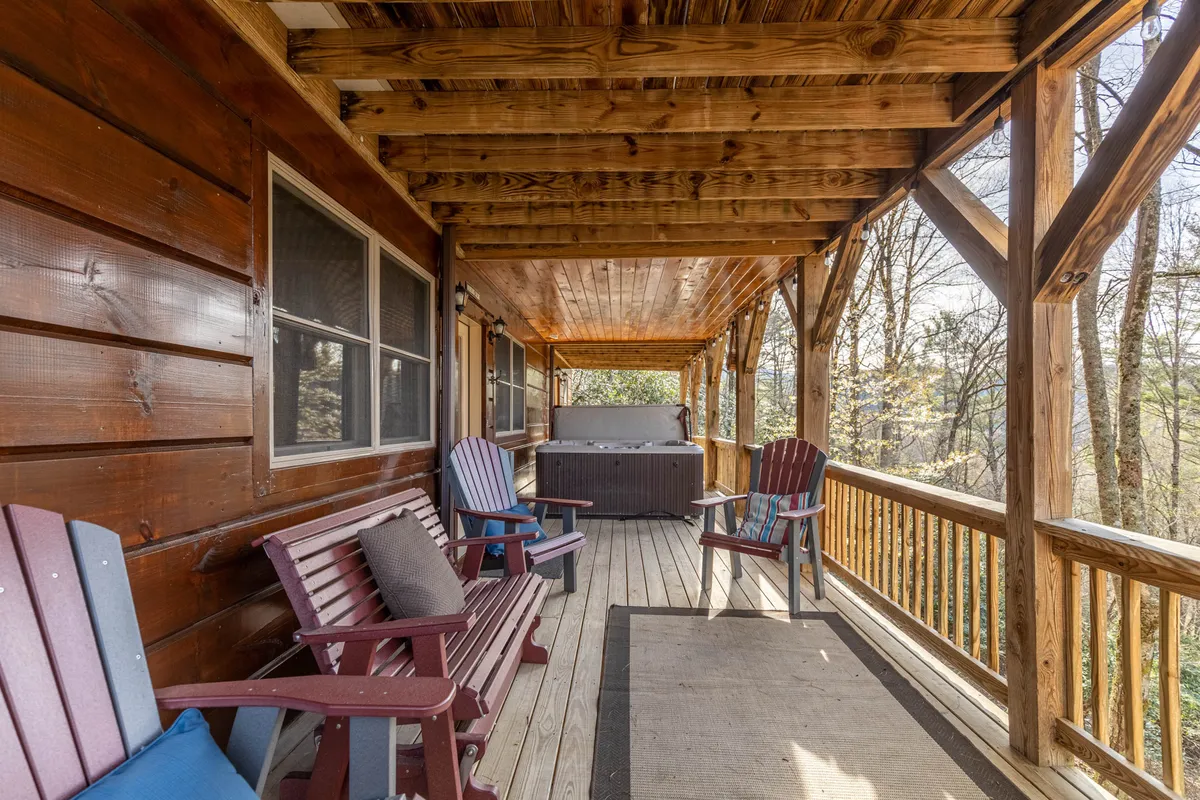An inviting lower deck featuring a bubbling hot tub, Adirondack chairs, and a rug, all surrounded by lush trees. The setup encourages outdoor relaxation, socializing, and soaking in the natural setting, with opportunities to gather around the fire pit nearby.