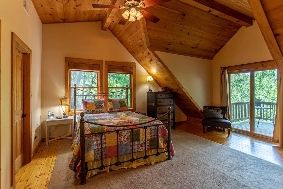 A serene bedroom featuring a bed near large windows, an armchair, and a dresser. The windows provide peaceful views of the outdoors, inviting relaxation and contemplation.