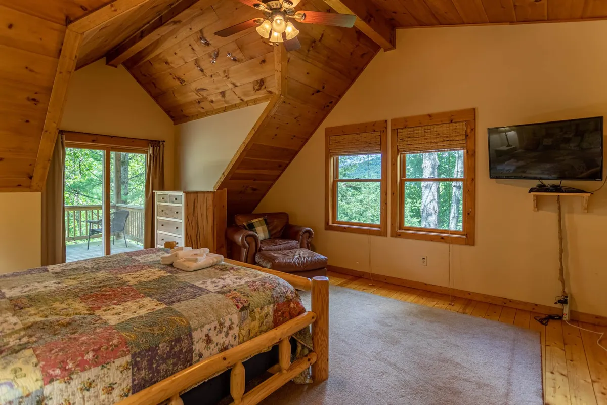 A tranquil bedroom featuring a bed with a patchwork quilt, large windows allowing plenty of natural light, wooden furniture, a leather armchair, and a smart TV mounted on the wall. The view from the room offers calming glimpses of the outdoors, creating a restful and inviting atmosphere.