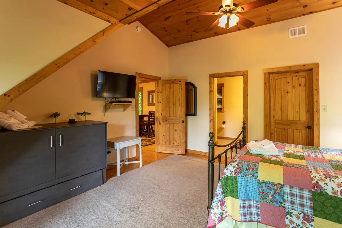 A comfortable bedroom setup featuring a quilted bedspread, wooden furniture, television, and open doors that suggest easy access and flow between spaces. The room is infused with warm lighting, fostering an atmosphere of coziness and relaxation.