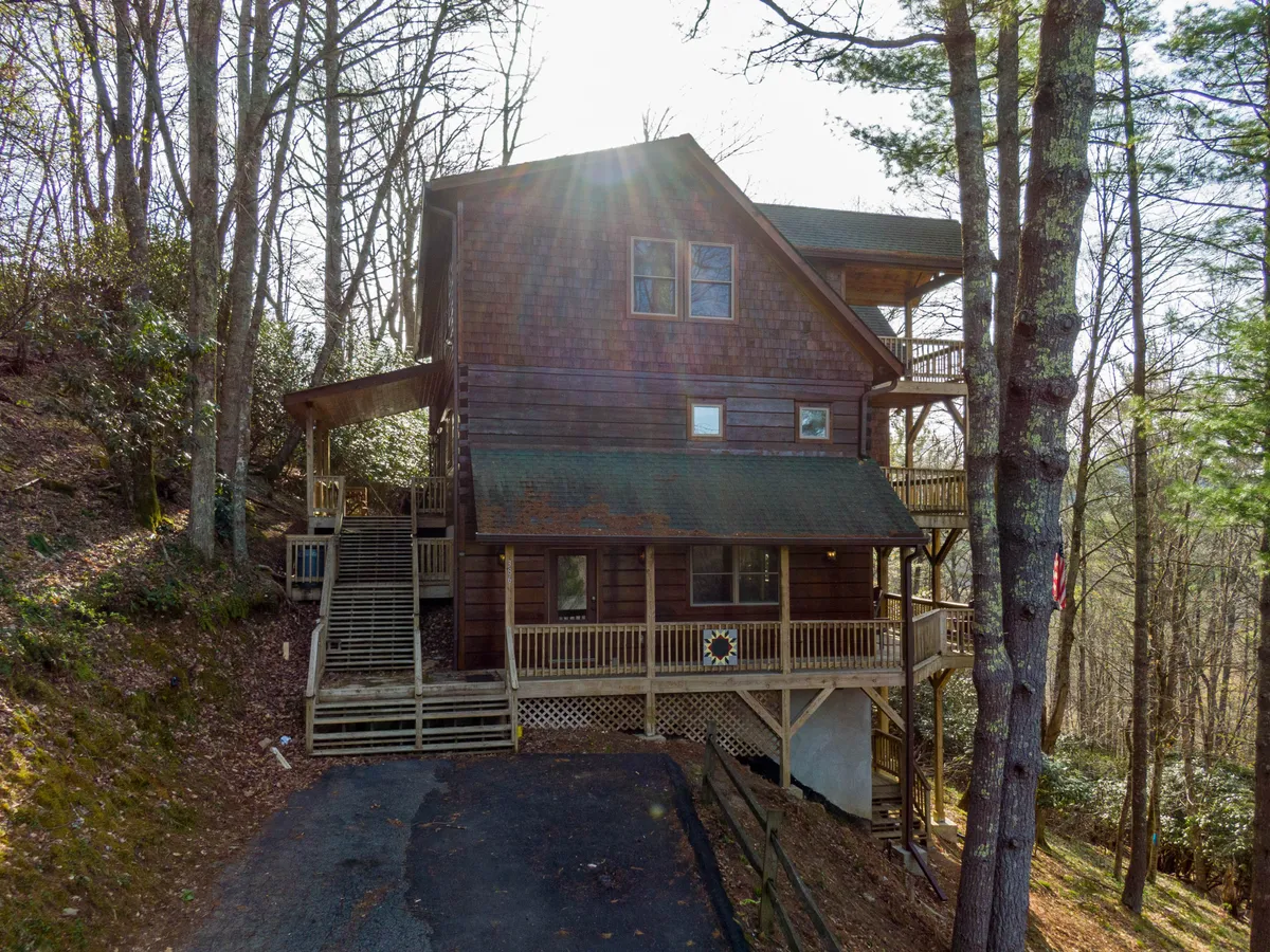 A wide wooden deck extends out into the trees, with stairs leading up and large windows connecting the indoor and outdoor spaces. The area is enveloped by nature and offers ample space for socializing, relaxing, or enjoying a morning coffee.