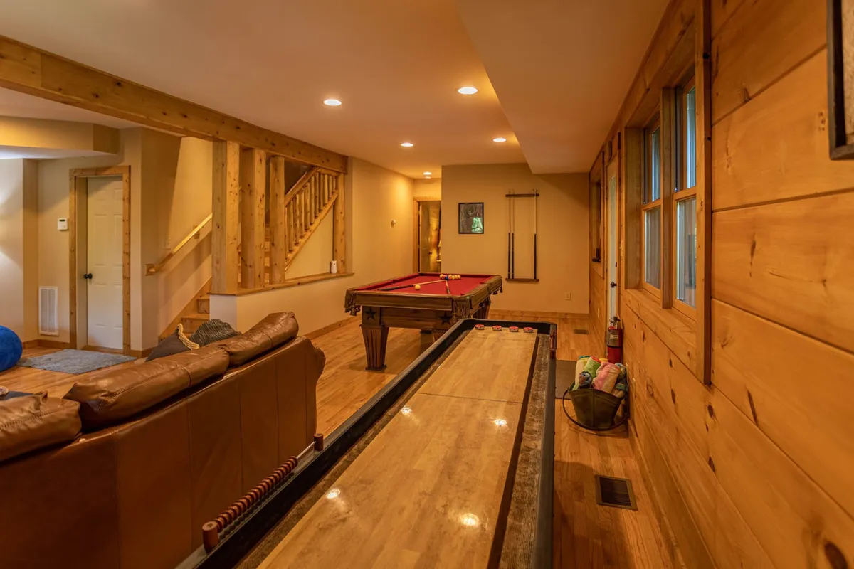 This image showcases a lower-level game room featuring a pool table and a shuffleboard table. The space is lined with wooden walls and includes a comfortable leather sofa, creating an inviting environment for socializing, friendly competition, and relaxation.
