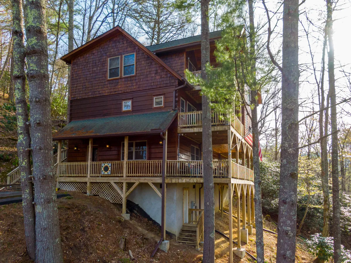 A rustic wooden porch with a railing and American flag overlooks the forested surroundings. The welcoming structure and spacious porch evoke feelings of arrival, outdoor gatherings, and the start of mountain adventures.
